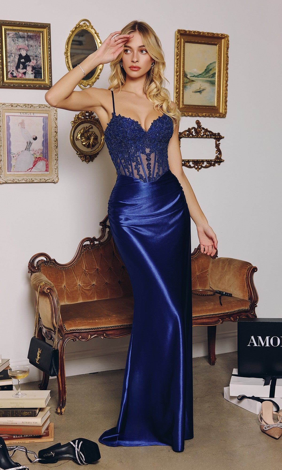 A woman in a fitted navy Long Prom Dress: Amoris T819 with an embellished corset bodice stands before a vintage sofa, surrounded by framed art, shoes, and shopping bags.